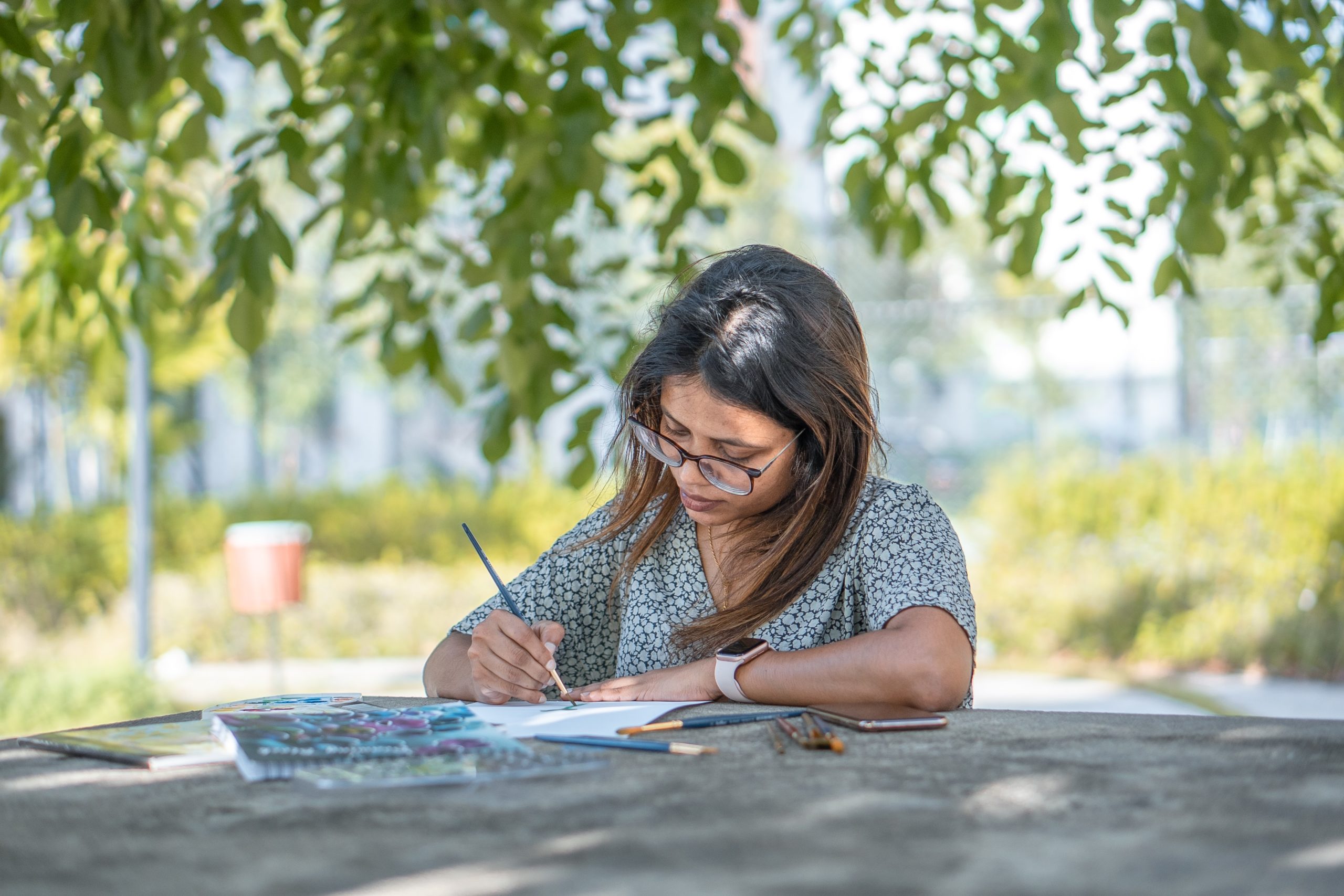 A woman with dark hair and glasses looks closely at the open book in front of her, pen poised to write. She is sitting outside and is surrounded by green leaves.