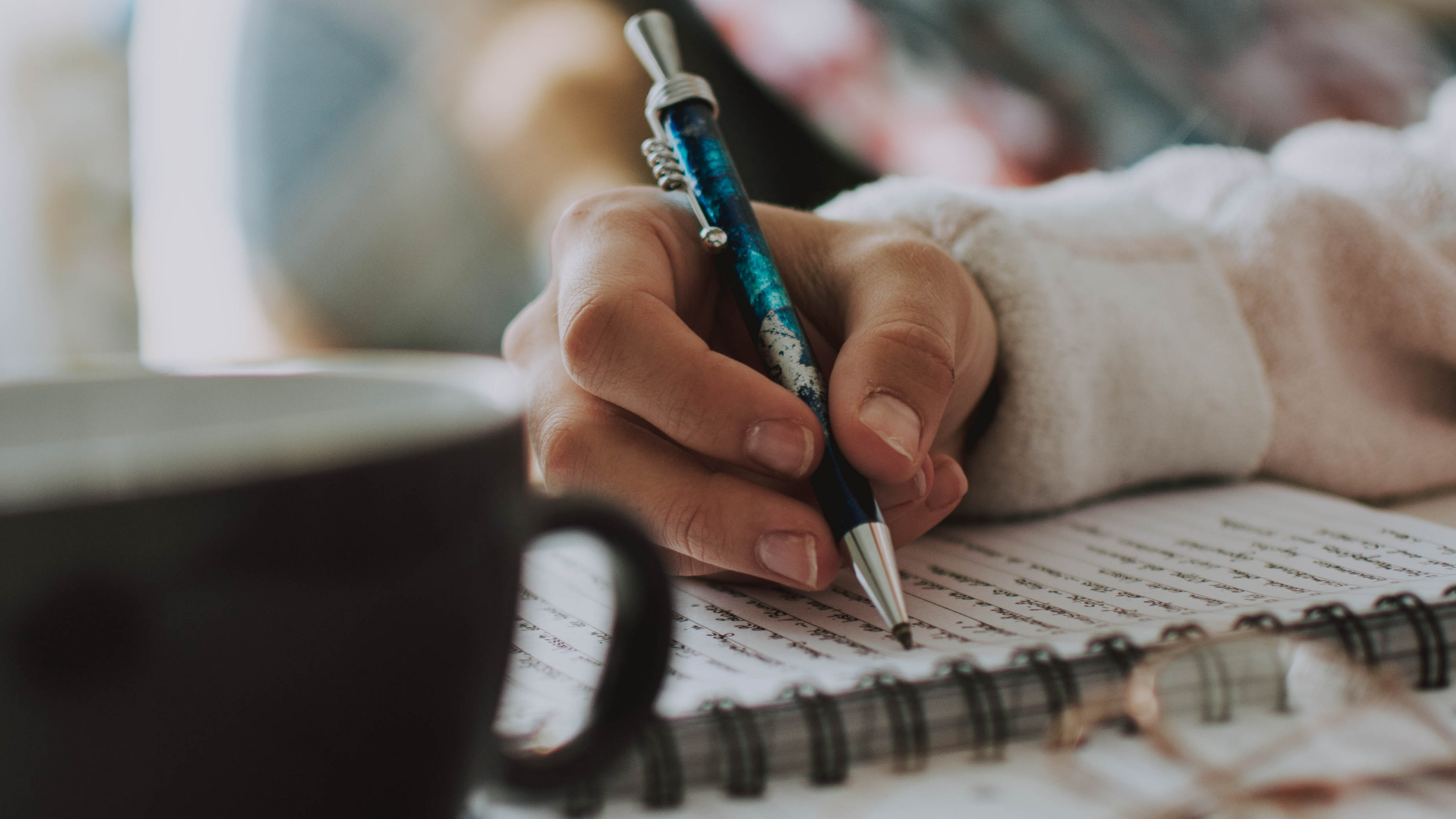 a hand is poised over a notebook, ready to write, in the foreground a black mug