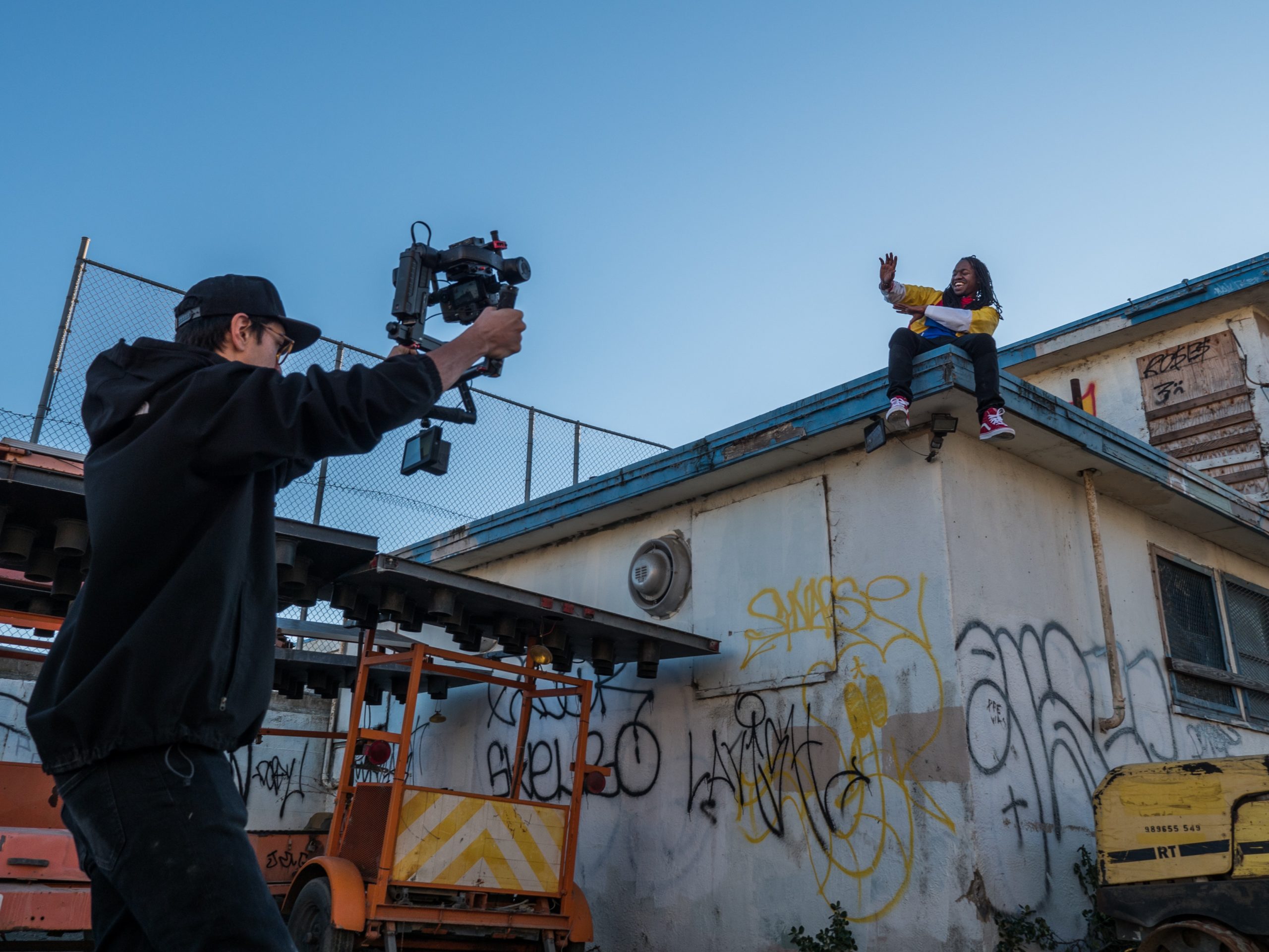 a dark man holds up a movie camera to film an actor sitting on the edge of a graffiti covered building
